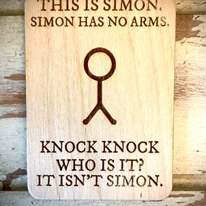 Puede incluir: Un letrero de madera con el texto "This is Simon. Simon has no arms." y un dibujo de un palo sin brazos. El letrero también tiene el texto "Knock knock who is it? It isn't Simon."