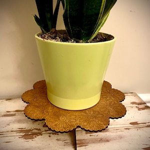 May include: A light green ceramic flower pot with a snake plant, on a wooden plant stand. The plant stand is flower-shaped with a floral pattern and three legs. The stand is made of wood. The pot and stand are on a distressed wooden surface.