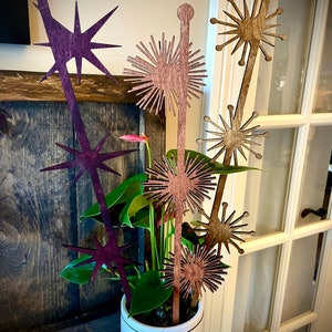 May include: A white ceramic planter with a green plant and five wooden decorative sticks. The sticks are shaped like stars and flowers. The flowers are brown and the stars are purple.