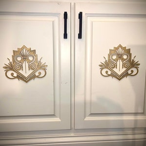 Może przedstawiać: Dwie białe drzwi szafki z czarnymi uchwytami i złotymi ozdobnymi akcentami. Akcenty są w stylu art deco.