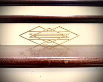 May include: A gold-colored geometric wall decor piece with a diamond shape, featuring a symmetrical design. The decor is mounted on a light-colored wall above a dark wooden shelf. The image also shows two dark wooden rails.