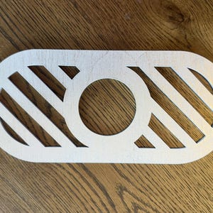 May include: A light-colored, oval-shaped wooden cutout featuring a central circular opening flanked by two sections of diagonal, parallel slats. The piece has a smooth, flat surface and a clean, laser-cut design with visible wood grain texture.
