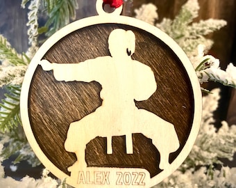 May include: A round wooden ornament with a silhouette of a person in a karate pose. The ornament is brown with a light wood finish and has the text "ALEX 2022" at the bottom.