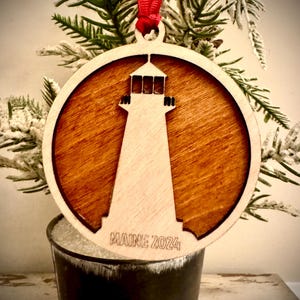 May include: A wooden Christmas ornament featuring a silhouette of a lighthouse with the text "MAINE 2024" underneath. The ornament is round and has a brown wood grain finish.