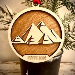 Pode incluir: Um enfeite de Natal de madeira com um design de pirâmides do Egito cortado a laser. O enfeite é marrom com um acabamento de madeira clara e tem o texto "EGITO 2024" gravado na parte inferior.