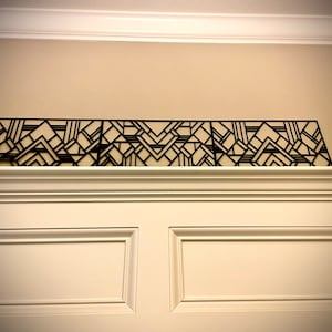 May include: A black geometric design wall decor made of metal. The design is made up of repeating patterns of squares and triangles. The decor is mounted on a white wall.