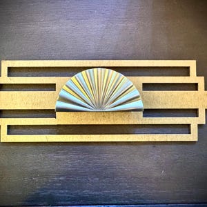 May include: A gold-colored metal drawer pull with a geometric design. The pull features a fan-shaped, silver-colored metal knob in the center.