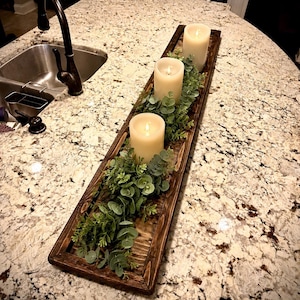 May include: A long, rectangular dark brown wooden tray containing three off-white pillar candles. The candles are surrounded by artificial green eucalyptus foliage and small leafy sprigs arranged along the length of the wooden base.