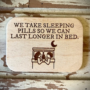 Puede incluir: Un letrero de madera con el texto "We take sleeping pills so we can last longer in bed." El letrero presenta una ilustración simple de una pareja en la cama con una luna creciente sobre ellos.