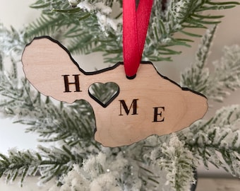 Puede incluir: Un adorno de madera con la forma de la isla de Maui, con la palabra "HOME" recortada. El adorno es de madera de color claro y cuelga de una cinta roja. Se exhibe en un pequeño árbol de Navidad nevado.