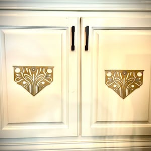 May include: Two white cabinet doors with black handles and gold decorative accents. The accents are intricate floral designs.