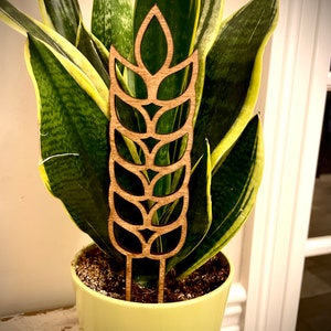 May include: A wooden plant stake shaped like a wheat stalk with a brown color. The stake is inserted into a green pot with a plant.
