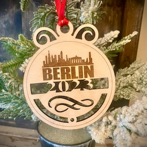 Könnte beinhalten: Ein hölzerner Weihnachtsbaumschmuck mit einem Laser-Cut-Design der Berliner Skyline und dem Text "BERLIN 2023". Der Schmuck hängt an einem roten Band.