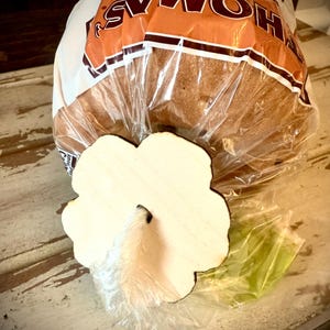 May include: A loaf of Thomas' Bagels in a clear plastic bag, with a wooden flower-shaped bagel holder on a distressed white surface. The bagel holder is light-colored with a hole in the center.