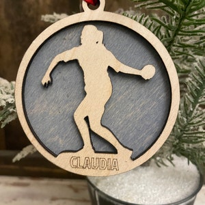 May include: A round wooden ornament with a silhouette of a person playing softball. The ornament is painted a dark gray color and has the name "CLAUDIA" engraved on the bottom.
