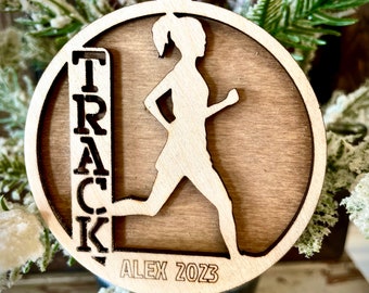 Peut inclure: Une décoration de Noël en bois représentant une silhouette de coureur avec le mot "TRACK" et le texte "ALEX 2023" sur un fond brun.
