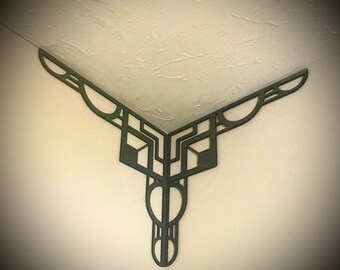 May include: A black metal corner bracket with a geometric design. The bracket is attached to a white wall.