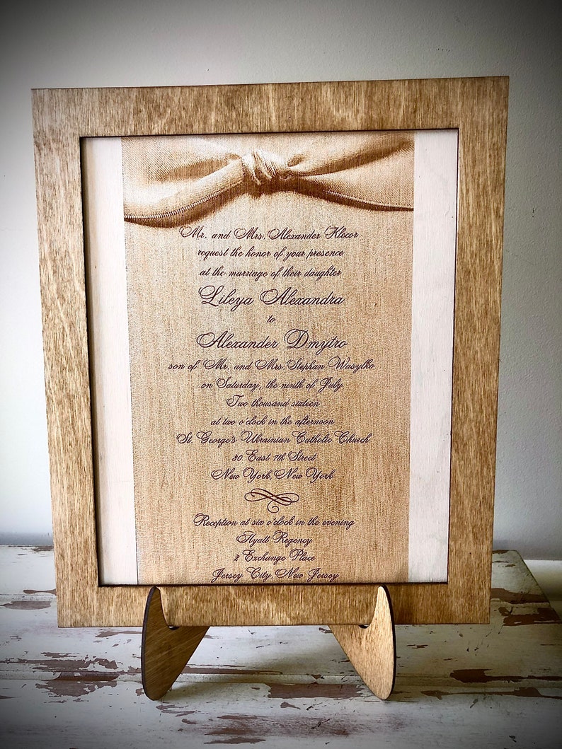 May include: A wedding invitation card with a brown wooden frame. The card is printed with a white background and brown text. The text reads: "Mr. and Mrs. Alexander Klecor request the honor of your presence at the marriage of their daughter Lileya Alexandra to Alexander Dmytro son of Mr. and Mrs. Stephan Wasylko on Saturday, the ninth of July Two thousand stateen at two o'clock in the afternoon St. George's Ukrainian Catholic Church 80 East 7th Street New York New York Reception at six o'clock in the evening Hyatt Regency 2 Exchange Place Jersey City, New Jersey"