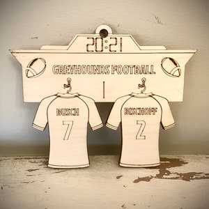 May include: A wooden sign with a scoreboard design featuring the text "GREYHOUNDS FOOTBALL" and the score "20:21". Two jerseys hang below the scoreboard, one with the name "BUSCH" and the number "7", and the other with the name "BISCHOFF" and the number "2".