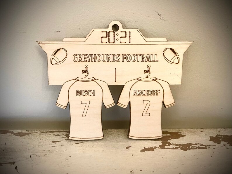 May include: A wooden sign with a scoreboard design featuring the text "GREYHOUNDS FOOTBALL" and the score "20:21". Two jerseys hang below the scoreboard, one with the name "BUSCH" and the number "7", and the other with the name "BISCHOFF" and the number "2".