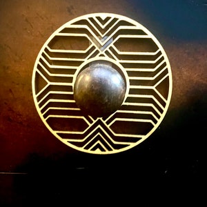 Könnte beinhalten: Ein runder, goldfarbener Metallknopf mit geometrischem Muster. Der Knopf hat eine dunkelbraune Mitte.