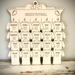 May include: A wooden sign with a football theme, featuring a scoreboard with the year 2021 and the text "Grizzly Football". The sign has 15 spaces for jerseys, each with a number and a name. The jerseys are all white with black outlines.