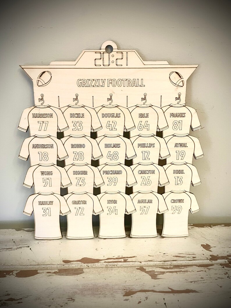May include: A wooden sign with a football theme, featuring a scoreboard with the year 2021 and the text "Grizzly Football". The sign has 15 spaces for jerseys, each with a number and a name. The jerseys are all white with black outlines.
