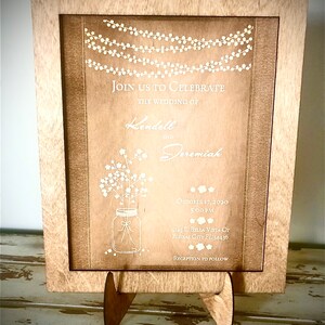 May include: A wooden framed wedding invitation with white text. The invitation reads "Join us to celebrate the wedding of Kendell and Jeremiah" with the date and time of the wedding, the address, and "Reception to follow".