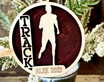 Peut inclure: Une décoration de Noël en bois avec une silhouette de coureur et le mot "TRACK" dans une bannière verticale. La décoration est peinte en rouge foncé et porte le texte "ALEX 2023" en bas.