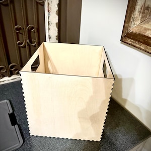 May include: A square, light-coloured wooden storage box with cut-out handles. The box has a natural wood grain finish and is constructed with interlocking edges. The box is sitting on a dark countertop.