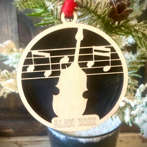 May include: A wooden Christmas ornament with a black background and a silhouette of a bass guitar with musical notes. The ornament is personalized with the name "ALEX" and the year "2022".