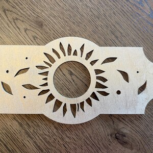 May include: A light-colored, flat wooden cutout featuring a central circular sun motif with radiating rays. The rectangular piece has decorative scalloped edges and symmetrical leaf-shaped cutouts on both sides, along with small circular holes.