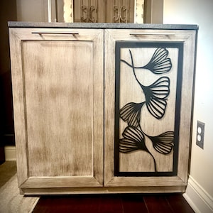 Puede incluir: Un armario de madera blanco con dos puertas. La puerta derecha tiene un recorte de metal negro de hojas de ginkgo. El armario tiene una encimera de granito y manijas plateadas.