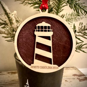 May include: A wooden Christmas ornament featuring a silhouette of a lighthouse with a red background. The ornament is round and has the text "NORTH CAROLINA 2024" inscribed on the bottom.