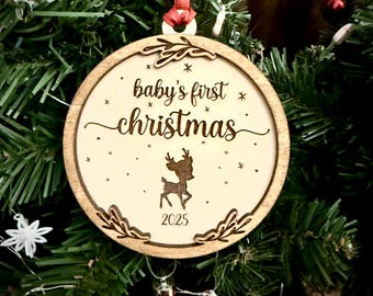 Op de afbeelding: Een houten kerstornament met de tekst "baby's first christmas" en het jaar 2025. Het ornament heeft een rendierontwerp en hangt aan een rood lint. Het ornament wordt omringd door groene kerstboomtakken en rode ornamenten.