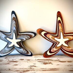 May include: Two wooden starfish wall decor, one in shades of gray and brown, the other in shades of brown and red. Both have a white starfish design in the center.