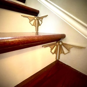 Może przedstawiać: Dwa złote metalowe akcenty dekoracyjne w geometrycznym wzorze są przymocowane do krawędzi drewnianych schodów. Akcenty mają kształt stylizowanego słońca.