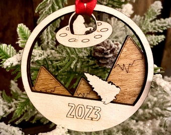 May include: A wooden Christmas ornament with a silhouette of a UFO and mountains. The ornament is decorated with a red ribbon and the year 2023 is engraved on the bottom.