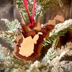 May include: A layered wooden ornament in the shape of West Virginia, with a red ribbon for hanging. The ornament features varying shades of brown wood, with a lighter wood center. The ornament is displayed against a backdrop of frosted evergreen branches.