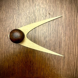 Könnte beinhalten: Ein goldfarbener Türgriff mit einem abgerundeten, dunkelbraunen Knopf. Der Griff ist boomerangförmig und auf einer Holzoberfläche montiert.