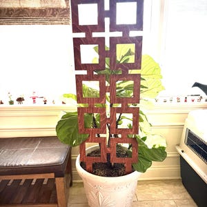 May include: A wooden plant trellis with a geometric square design in a white decorative pot. The trellis is a dark reddish-brown color and is placed in front of a large-leafed plant. The pot has a floral pattern.