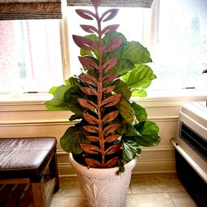 May include: A wooden plant stake in a leaf design is placed in a white decorative pot with a large green plant. The stake is a reddish-brown color and has a detailed leaf pattern. The pot has a floral design.