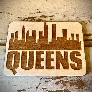 May include: A wooden coaster with a brown silhouette of the New York City skyline and the word "QUEENS" in brown lettering.