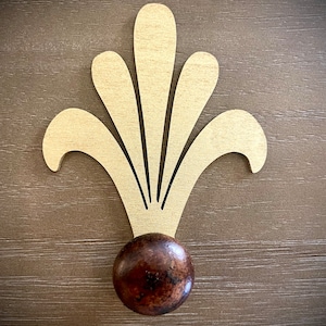 May include: A wooden wall hook with a decorative, stylized floral design in a light brown color. The hook has a round, dark brown wooden knob.