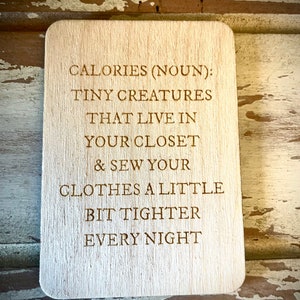 May include: A wooden sign with the text "CALORIES (NOUN): TINY CREATURES THAT LIVE IN YOUR CLOSET & SEW YOUR CLOTHES A LITTLE BIT TIGHTER EVERY NIGHT".