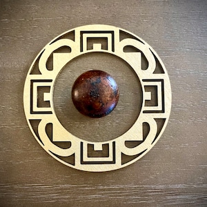 May include: A wooden knob with a geometric design in a gold color. The knob is round and has a brown wooden center.