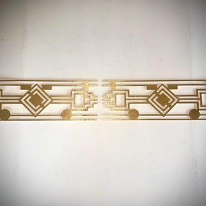 May include: Two Art Deco-style gold-colored metal wall art pieces. Each piece features geometric shapes, including squares, diamonds, and circles, arranged in a symmetrical pattern. The design is reminiscent of the 1920s and 1930s.