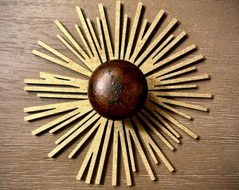 Könnte beinhalten: Ein goldfarbenes Sonnenstrahlen-Design mit einem braunen Holzknauf in der Mitte. Der Sonnenstrahlen-Effekt besteht aus dünnen, flachen Metallstücken, die sich vom Zentrum nach außen erstrecken.