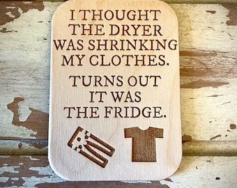 May include: Wooden sign with a humorous saying about a dryer shrinking clothes. The text reads "I thought the dryer was shrinking my clothes. Turns out it was the fridge." The sign has a graphic of a shirt and a pair of pants.