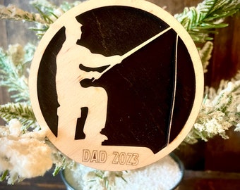 May include: A wooden Christmas ornament with a silhouette of a person fishing. The ornament is painted black and has the text "DAD 2023" engraved on the bottom.